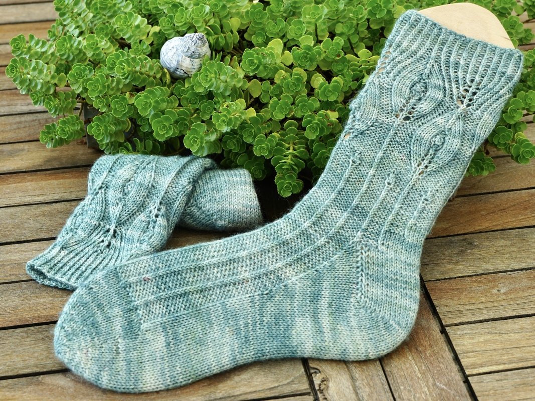 Paar hellgrün-melierter gestrickter Socken mit Blattmuster auf Holzbrett vor grünen Sukkulenten