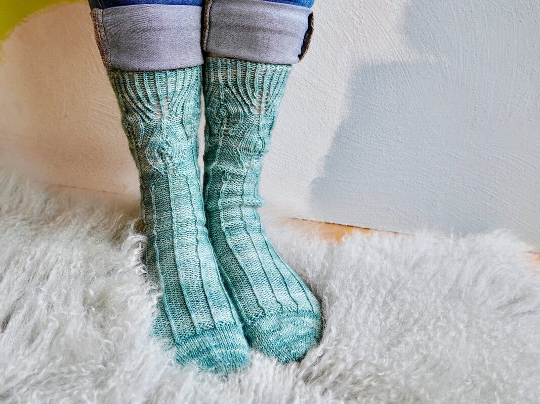 Feet wearing teal ribbed socks with cable knit pattern on a white shag rug.
