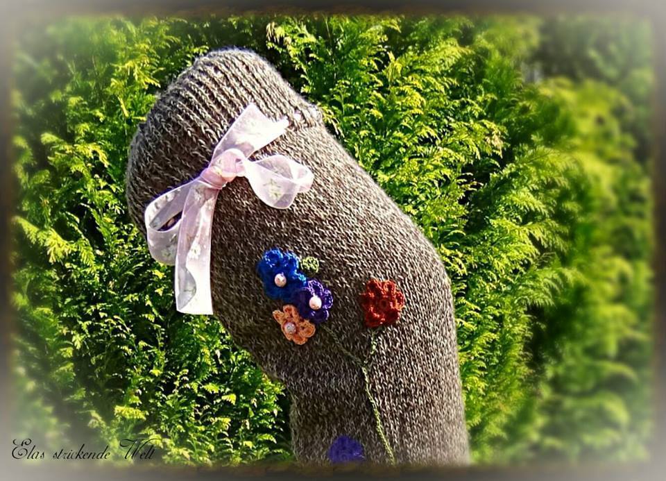 Gestrickte graubraune Socke mit rosa Schleife und blauen, orangefarbenen und roten Häkelblumen vor grünem Laub