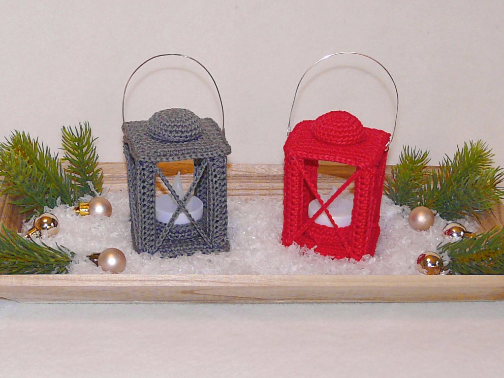 Two small crocheted lanterns in red and gray on a wooden tray with faux snow, pine sprigs, baubles and tea lights