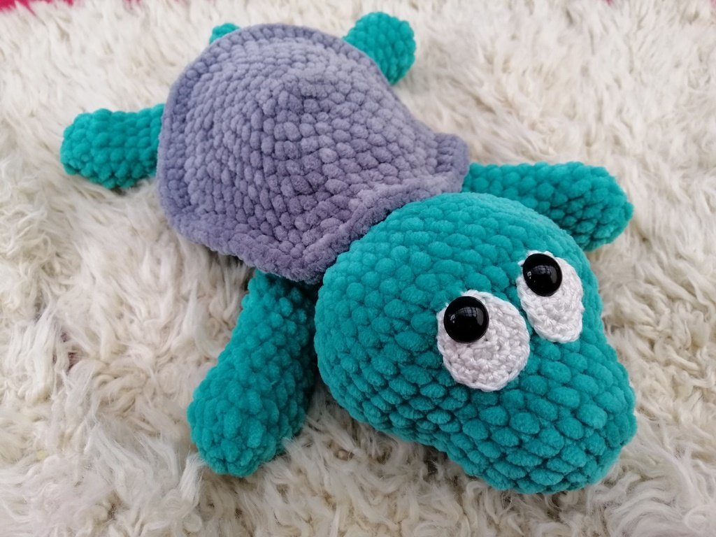 Türkis‑graue gehäkelte Amigurumi‑Schildkröte mit großen Augen auf hellem Fell