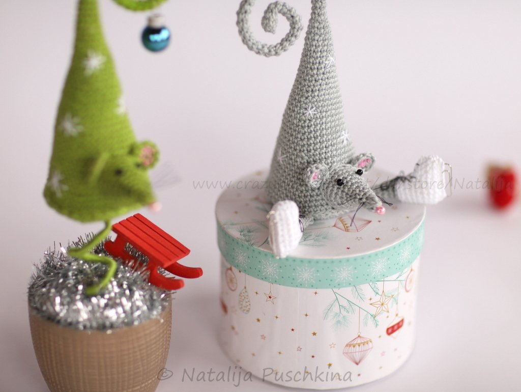 Zwei gehäkelte Mäuse in Tannenbaumform, eine graue auf einer Geschenkbox, eine grüne mit Mini-Schlitten und Lametta.