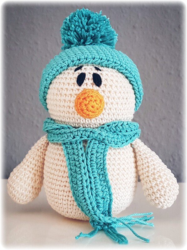 Gehäkelter weißer Schneemann mit türkisfarbener Bommelmütze, passendem Schal, orangefarbener Nase und schwarzen Augen.