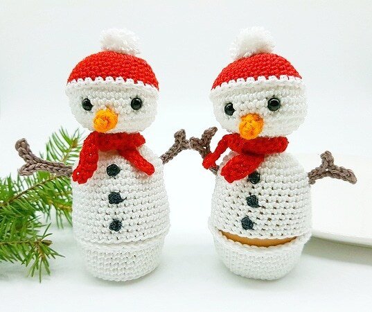 Zwei gehäkelte Schneemann-Eierwärmer mit roten Mützen und Schals. Einer der Schneemänner wärmt ein Ei. Beide haben orange Karottennasen, schwarze Knöpfe und braune Ärmchen.