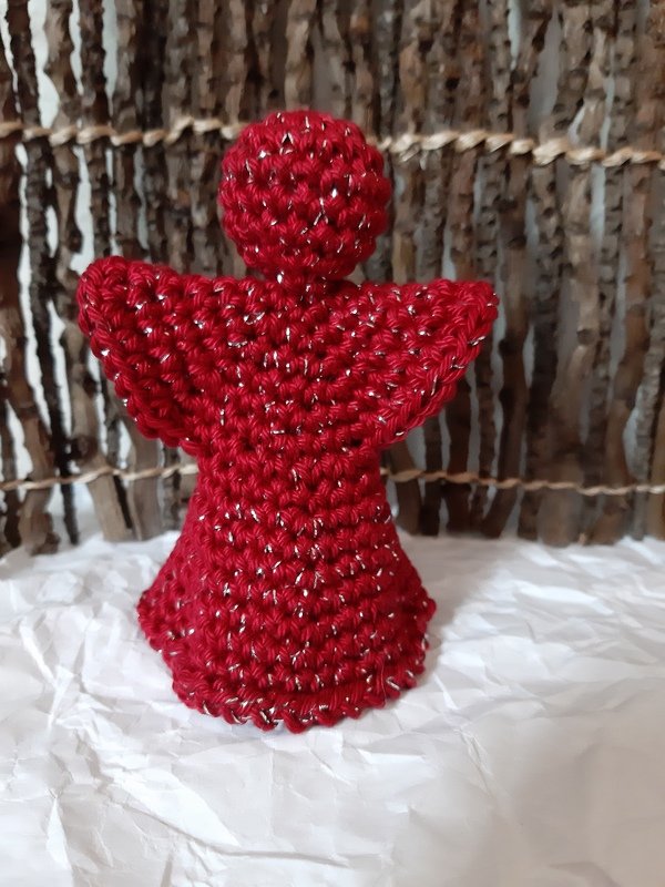 Gehäkelter roter Engel mit silbernem Glitzer vor rustikaler Holzwand auf weißem, zerknittertem Untergrund.
