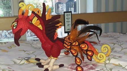 Gehäkelter Phönix‑Amigurumi in Rot, Orange und Gelbtönen mit spiralförmigem Schwanz und federähnlichen Verzierungen auf Stoffuntergrund