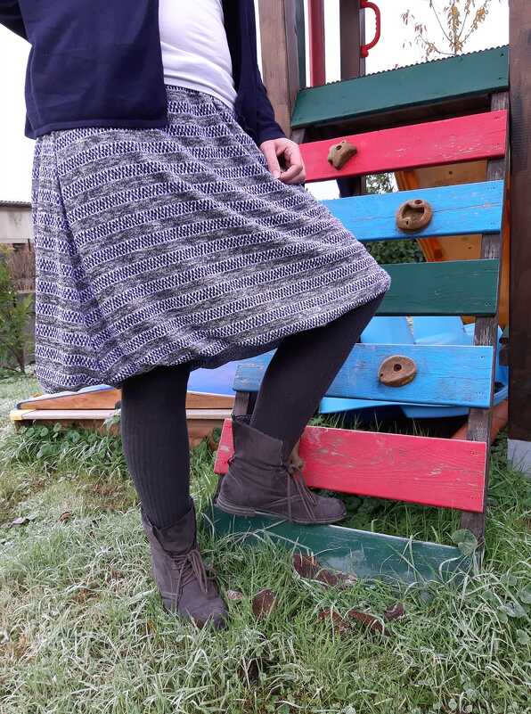 Frau mit grauem, gestreiftem Rock, dunklen Strumpfhosen und braunen Stiefeletten vor buntem Spielgerät