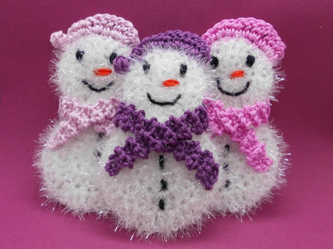 Drei gehäkelte Schneemänner aus schimmerndem Garn mit rosa- und lilafarbenen Mützen und Schals vor pinkem Hintergrund