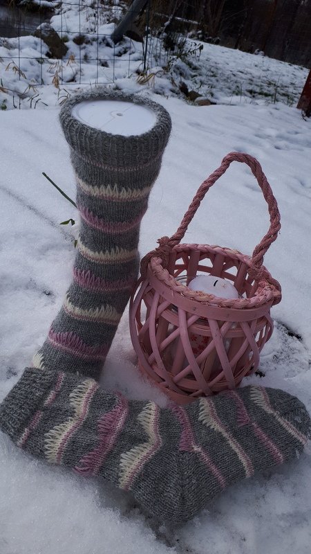 Nahaufnahme gestrickter Beinstulpe und Socke mit Rippenbündchen und Streifen in Grau, Rosa und Creme im Schnee neben rosa Laterne