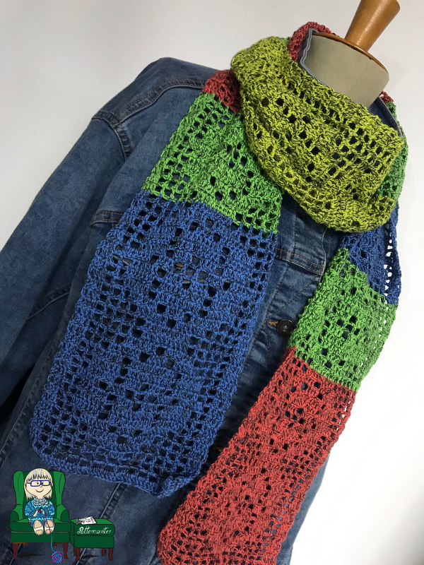 Gehäkelter Schal mit Gittermuster in Grün, Blau und Rot, locker über einer blauen Jeansjacke drapiert.