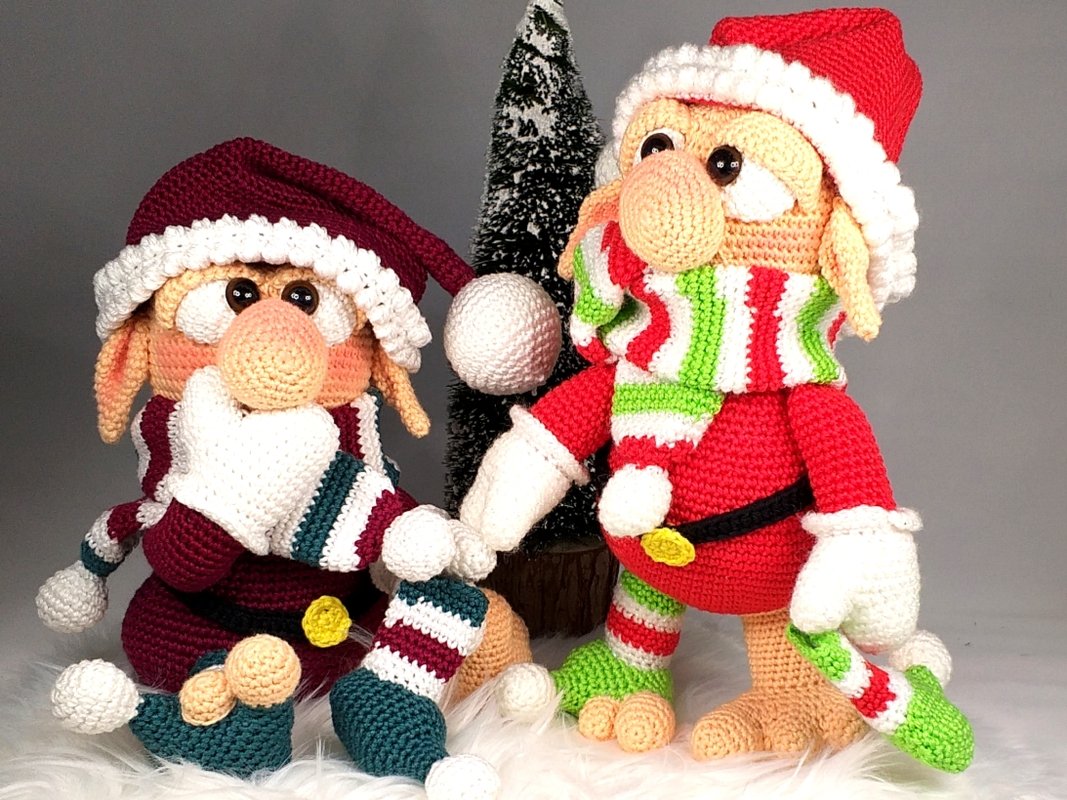 Two crocheted amigurumi gnomes in red and burgundy outfits with striped scarves and pom-pom hats beside a small tabletop tree