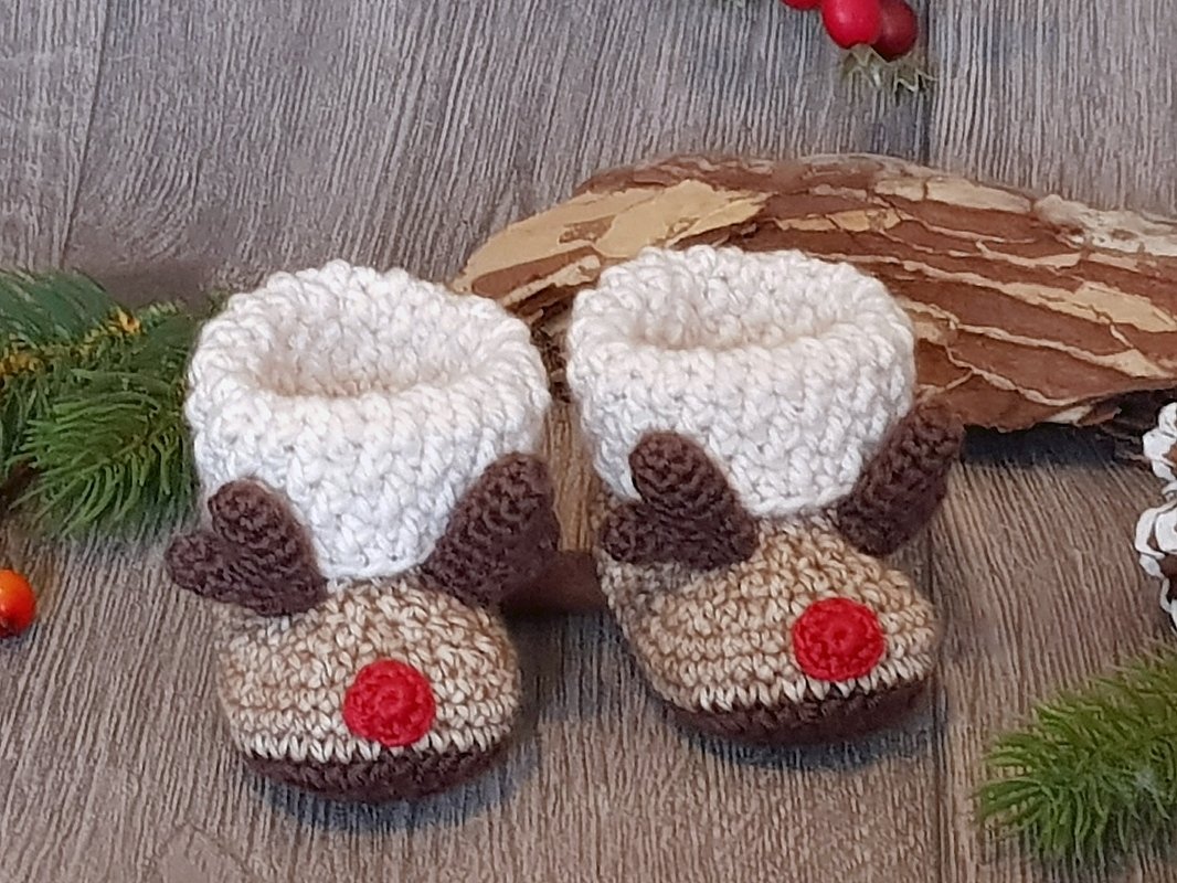 Pair of crocheted reindeer baby booties with cream cuffs, brown antlers and red noses on a wooden floor