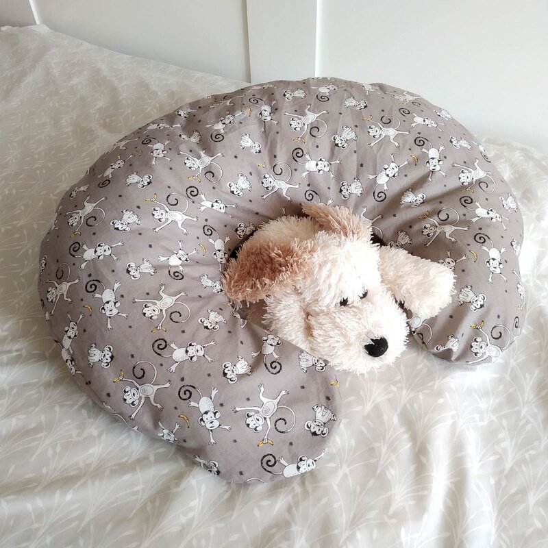Graues U‑förmiges Kissen mit Affenmuster und Plüschhund auf hellem Bett.