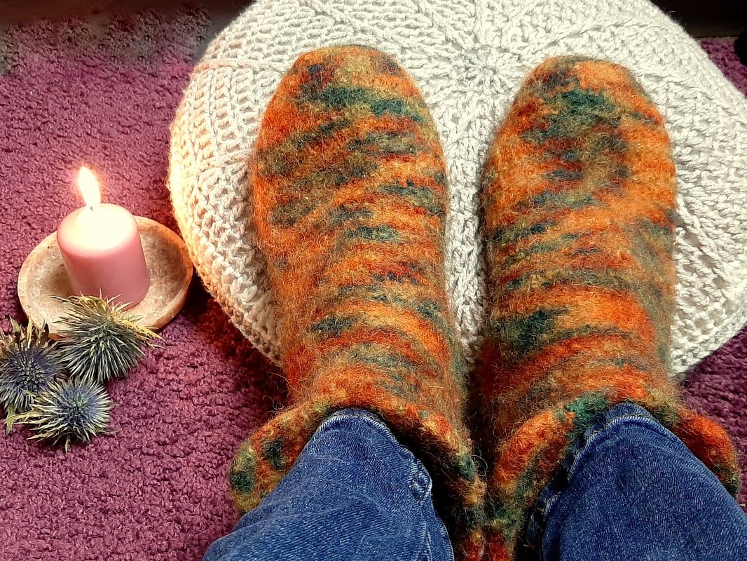 Orange‑grün‑braune Strickfilzpuschen auf gestricktem Kissen neben rosa Kerze und getrockneten Disteln auf violettem Teppich.