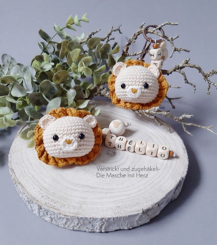 Zwei gehäkelte Löwenköpfe, einer als Schlüsselanhänger mit Holzbuchstabenperlen, der andere dekorativ auf einer Baumscheibe.