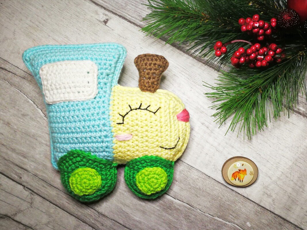 Gehäkelter Amigurumi-Zug in Blau, Gelb und Grün mit geschlossenem Gesicht auf hellem Holz, daneben Tannenzweige und rote Beeren.
