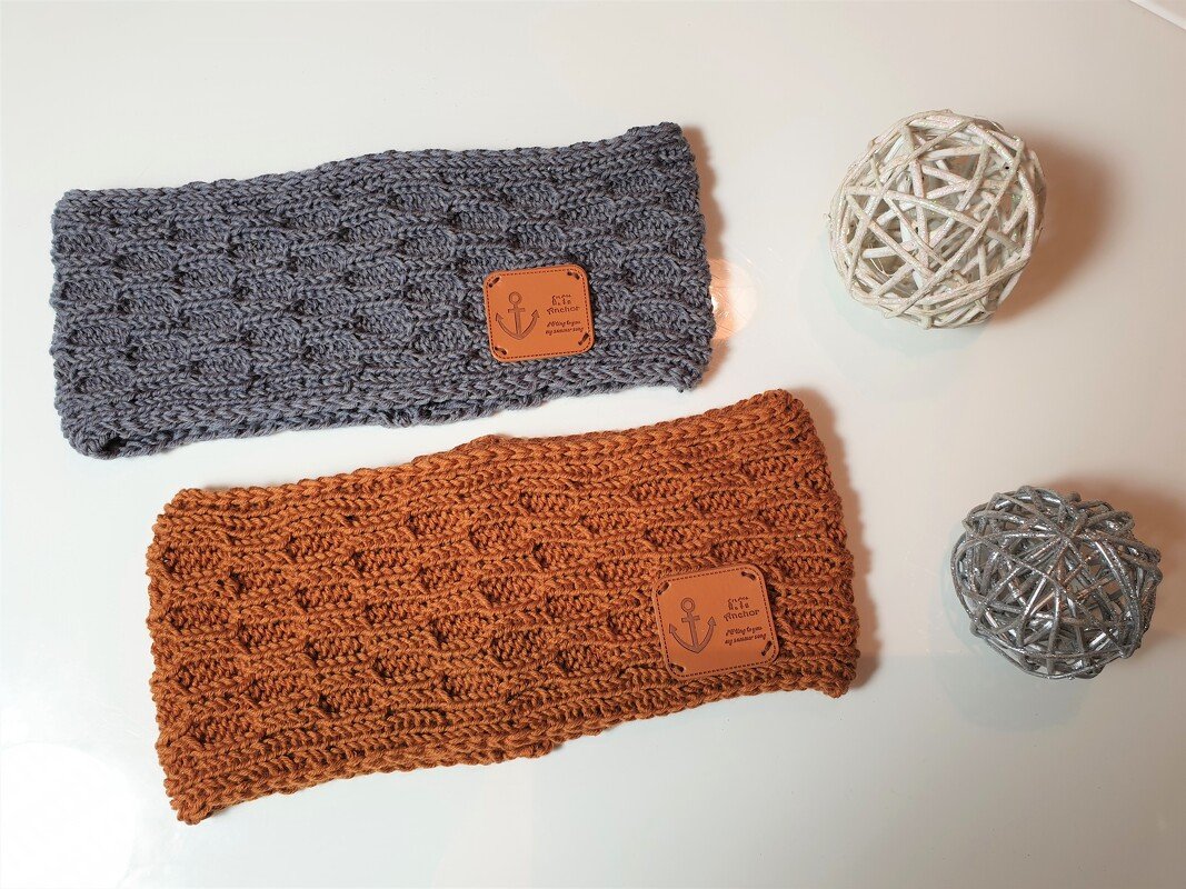 Zwei gestrickte Stirnbänder auf hellem Untergrund, eines grau, eines rostbraun, beide mit Lederetikett mit Anker‑Motiv.