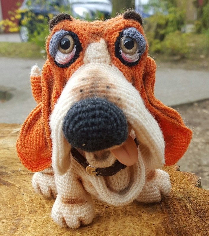 Gehäkelter Amigurumi Basset-Hund mit orange-braunen Schlappohren, großer schwarzer Nase, Halsband und herausgestreckter Zunge auf einem Holzstamm