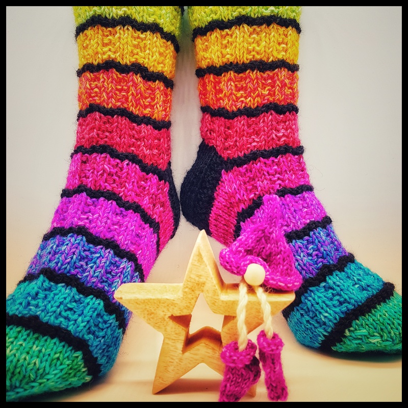 Nahaufnahme gestrickter mehrfarbiger Socken mit breiten Ringeln und schwarzen Trennlinien, davor Holzstern und kleines gestricktes Mützchen