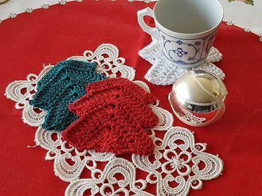 Gehäkelte rote und petrolfarbene Blatt-Untersetzer auf weißer Spitze neben Tasse und silberner Weihnachtskugel