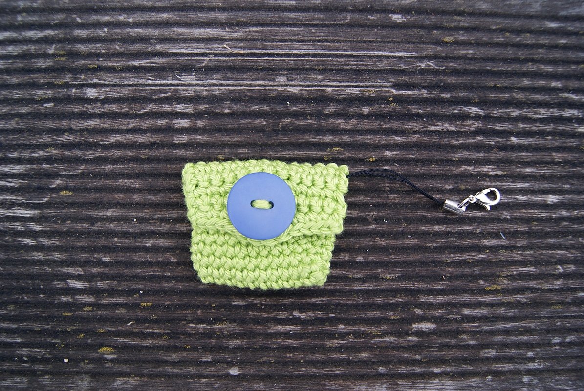 Hellgrünes gehäkeltes Täschchen mit blauem Knopf und schwarzem Band mit Karabiner auf dunklem Holzuntergrund