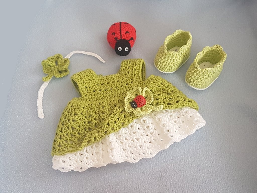 Hellgrünes und weißes gehäkeltes Babykleid mit Schühchen, Kleeblatt-Haarband und rotem Marienkäfer-Amigurumi auf hellgrauem Untergrund