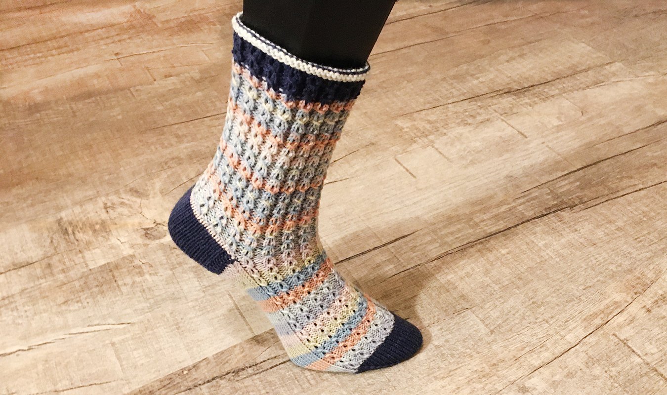 Mehrfarbiger gestrickter Strumpf mit Wabenmuster in Pastelltönen und dunkelblauen Akzenten auf einem Fuß auf Holzboden.