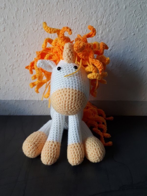 Gehäkeltes Amigurumi-Einhorn in Weiß mit oranger lockiger Mähne, sitzend