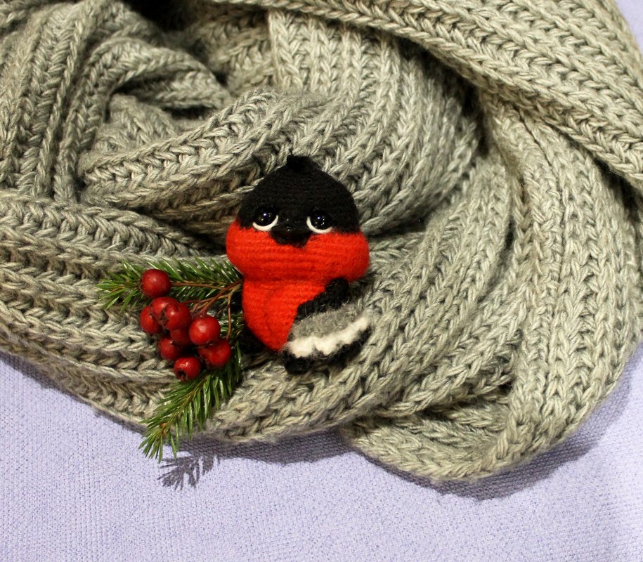 Gehäkelter Dompfaff auf grauem Strickschal mit Tannenzweig und roten Beeren