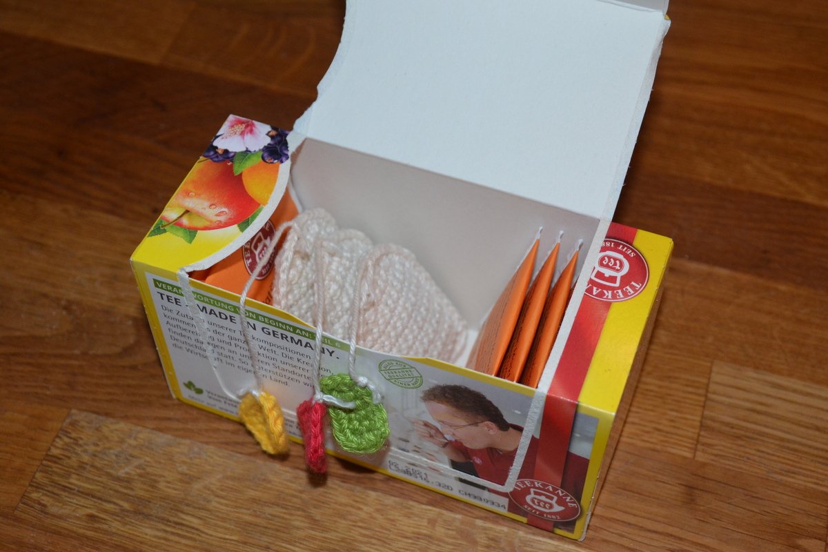 Offene Teebox mit Teebeuteln, hellen gehäkelten Untersetzern und bunten gehäkelten Anhängern