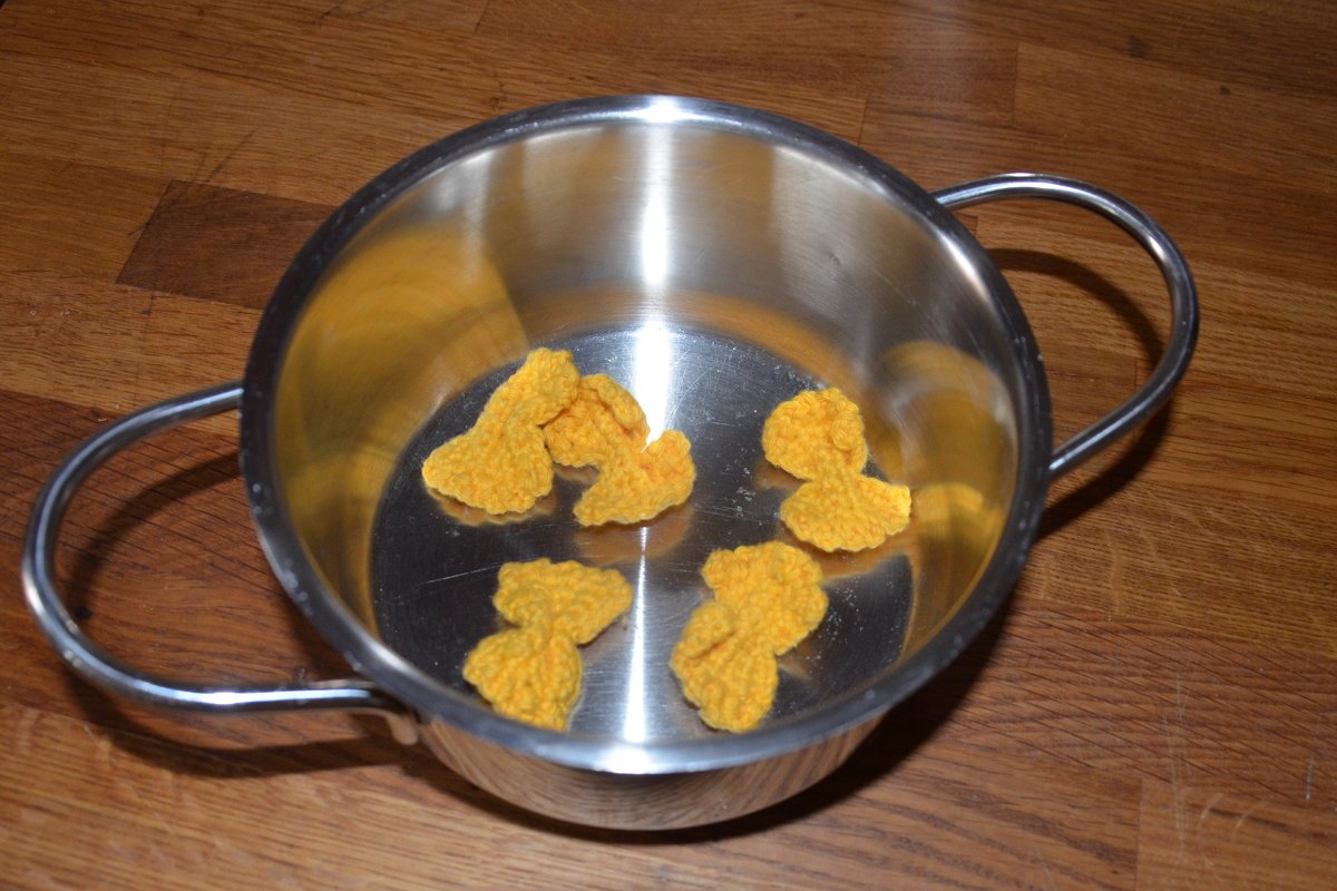 Fünf gelbe, gehäkelte Mini-Figuren in einem glänzenden Edelstahltopf auf Holz