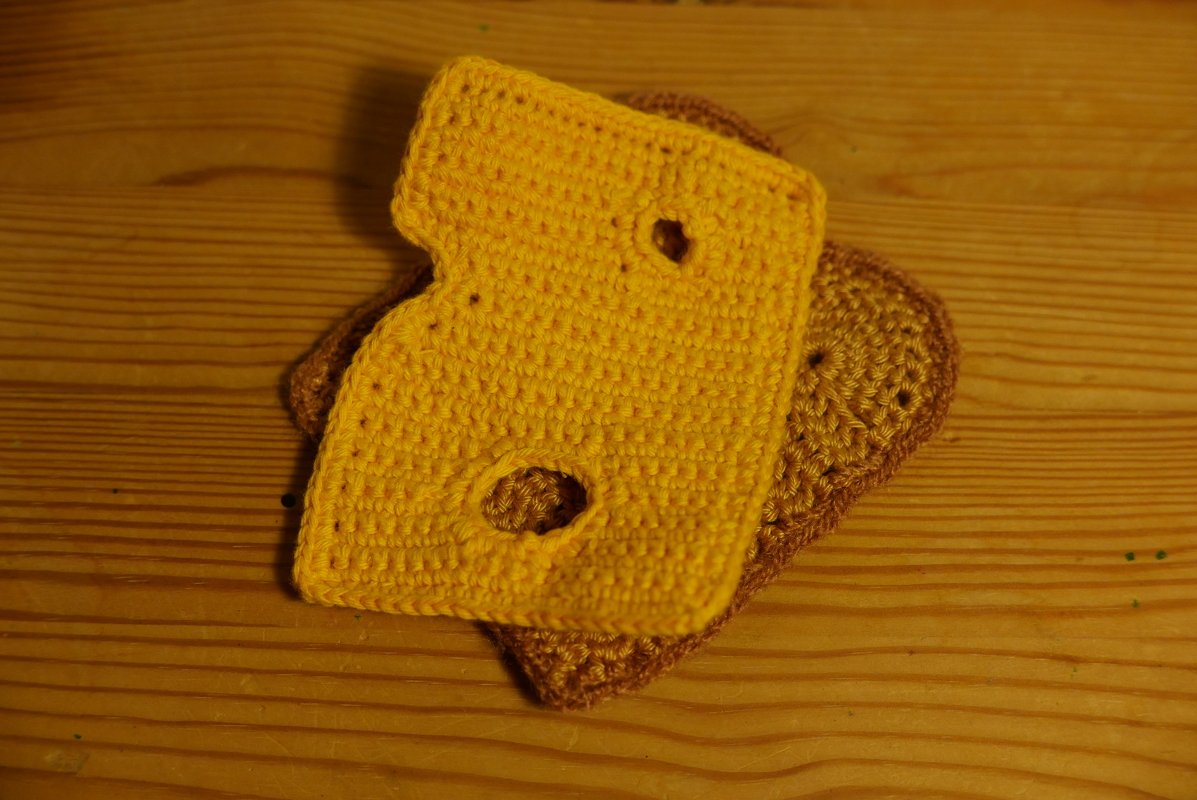 Gelbe gehäkelte Käsescheibe mit zwei Löchern auf braunem gehäkeltem Toast auf Holzoberfläche.