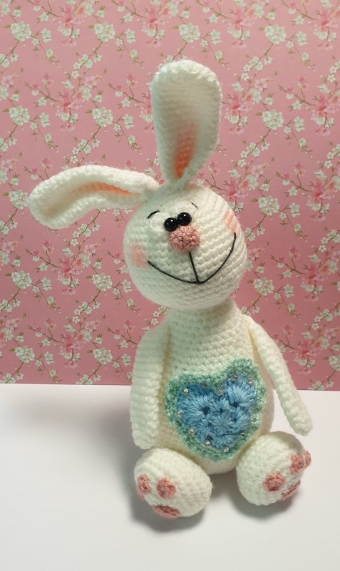 Weißer gehäkelter Amigurumi-Hase mit blauem Herz, rosa Pfoten und rosa Nase vor floralem Hintergrund.