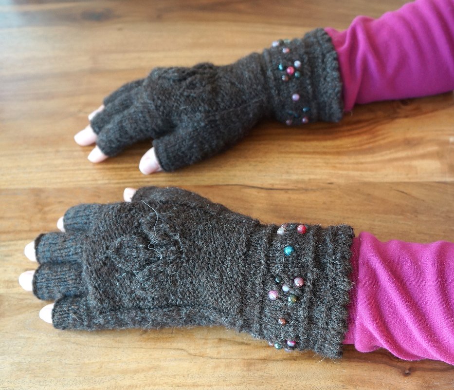 Braune gestrickte fingerlose Handstulpen mit Zopfmuster und bunten Perlen am Bündchen, getragen über rosa Ärmel auf Holzoberfläche