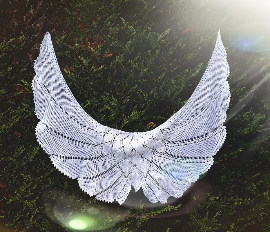 Weißes halbrundes Häkeltuch mit feinem Loch- und Spitzenmuster, aufgehängt vor dunkelgrünem Gebüsch, Lichtschein von rechts oben