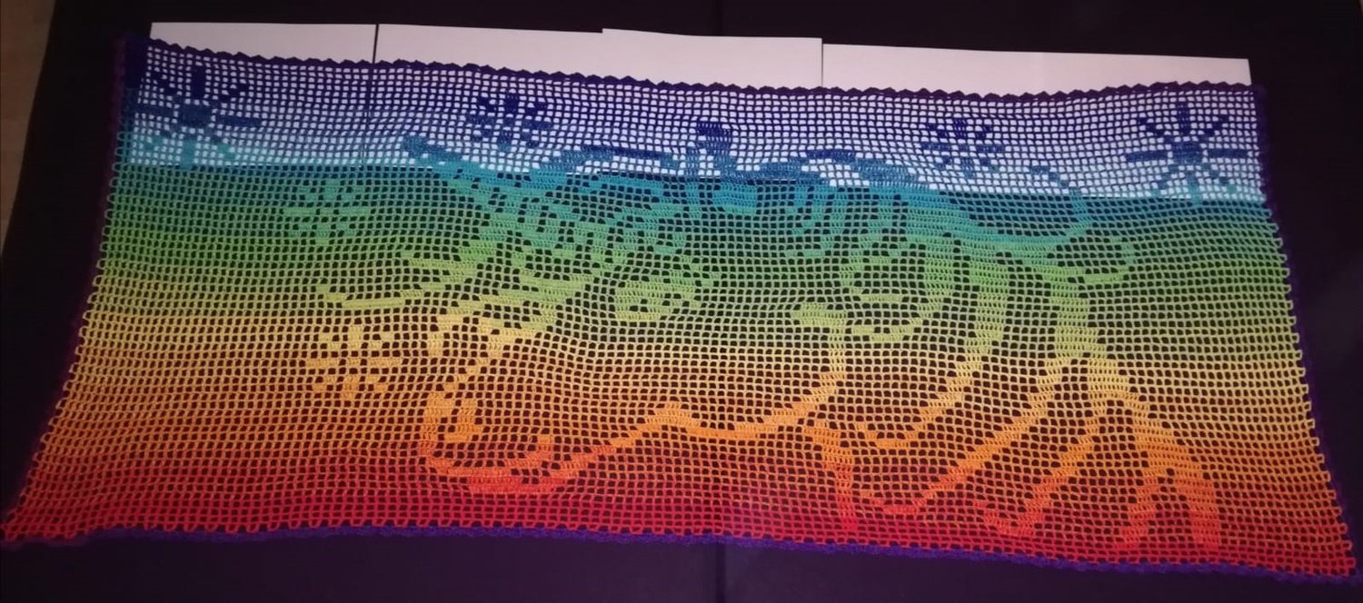 Gehäkeltes Filet-Tuch mit Regenbogen-Farbverlauf und durchbrochenen Stern- und Wellenmustern auf dunklem Hintergrund.