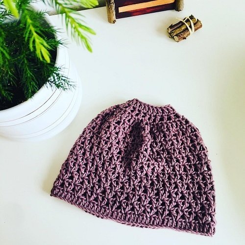 Lila gehäkelte Mütze mit Öffnung für Pferdeschwanz auf weißer Fläche neben Zimmerpflanze.