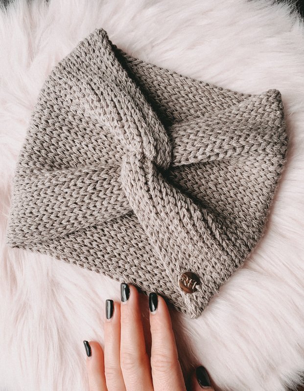 Grau gehäkeltes Stirnband mit Twistknoten auf hellem Fell, Hand mit schwarzen Nägeln berührt es