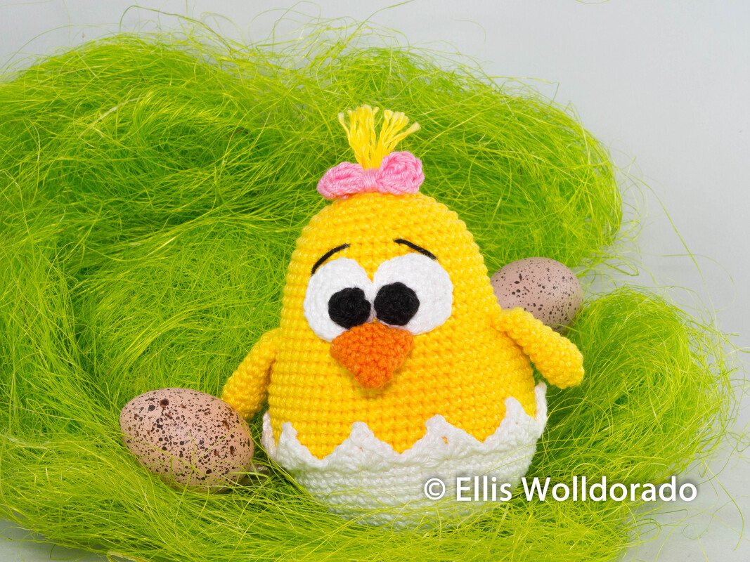 Gelbes Amigurumi-Küken mit rosa Schleife in weißer Eierschale auf grünem Dekogras, mit zwei gesprenkelten Ostereiern.