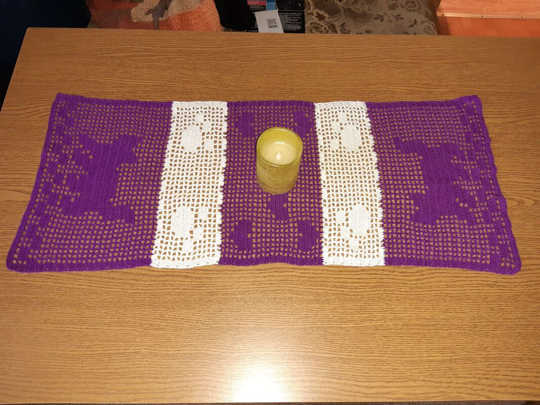 Lila und weiß gehäkelter Tischläufer auf einem Holztisch mit einer goldfarbenen Kerze in der Mitte.