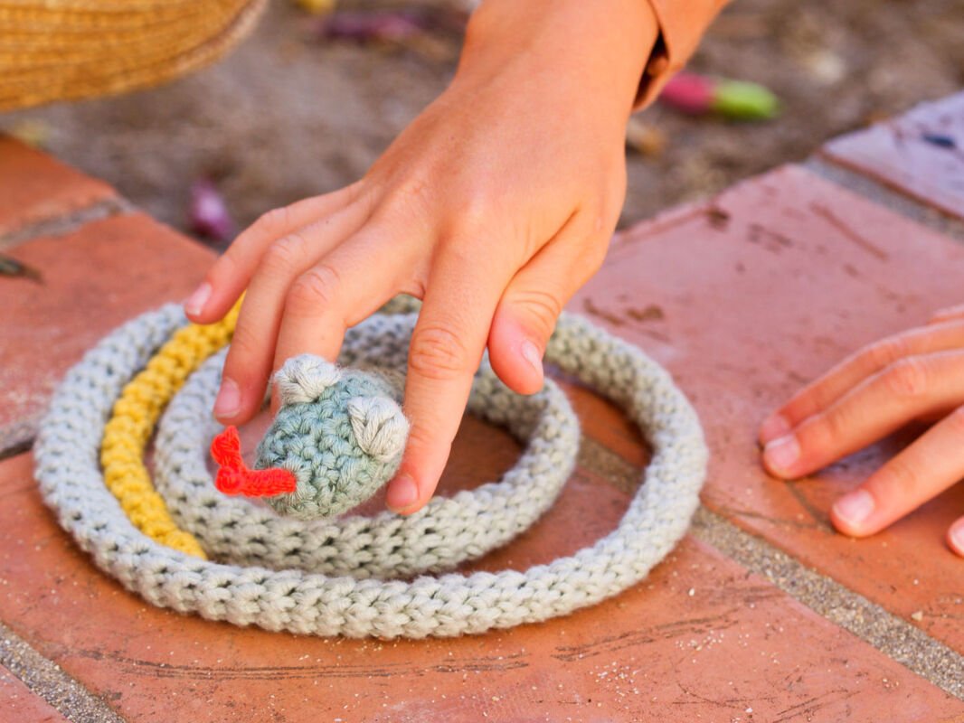 Gehäkelte geringelte Amigurumi-Schlange in Grau und Gelb mit roter Zunge, von einer Kinderhand berührt auf Ziegelboden.