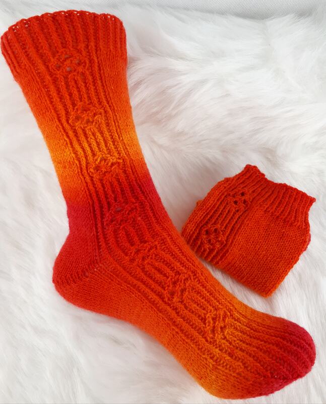 Orangene gestrickte Socke mit Farbverlauf und Zopfmuster neben passendem Bündchen