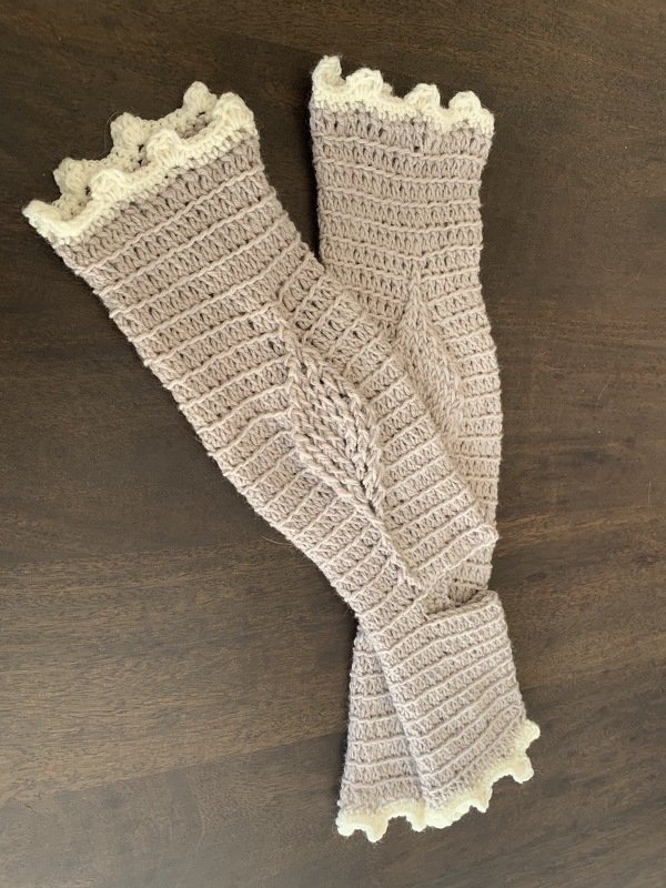 Paar hellbeige gehäkelte Beinstulpen mit cremefarbener Rüschenkante auf dunklem Holz