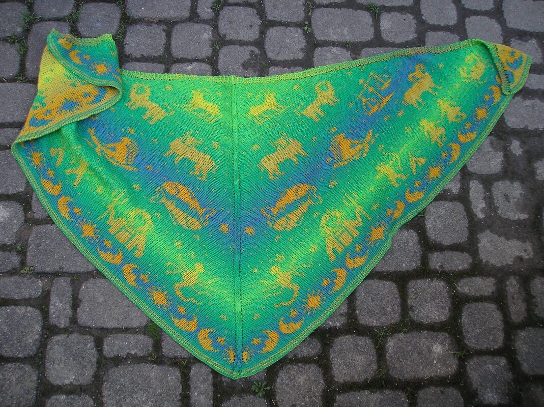 Gestricktes dreieckiges Tuch in Grün-Türkis mit gelb-orangenen Sternzeichen-Motiven liegend auf Kopfsteinpflaster