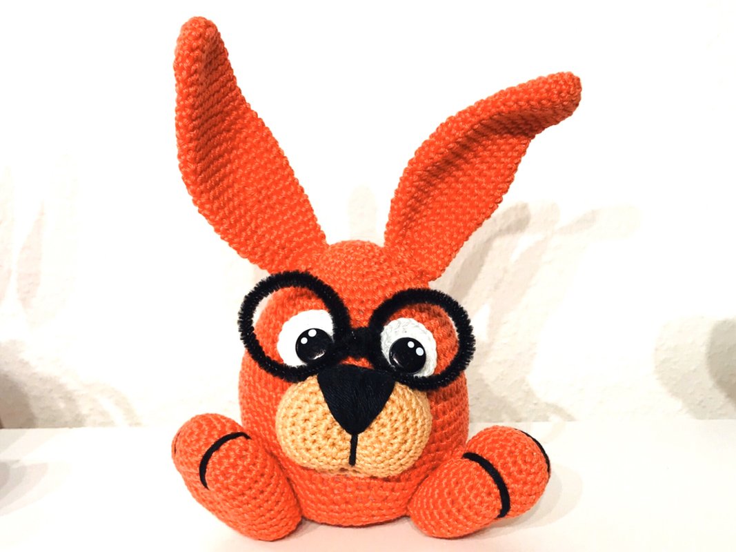 Gehäkelter oranger Hase mit runder schwarzer Brille und langen Ohren auf hellem Hintergrund.