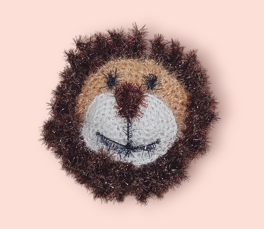 Gehäkelter Löwenkopf mit dunkelbrauner, buschiger Mähne und hellem Gesicht auf rosa Hintergrund