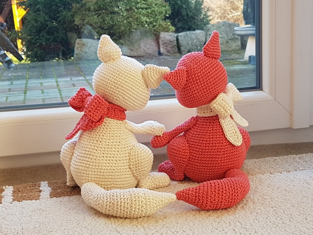 Zwei gehäkelte Katzen in Creme und Koralle mit Schals sitzen dicht nebeneinander auf einer Fensterbank