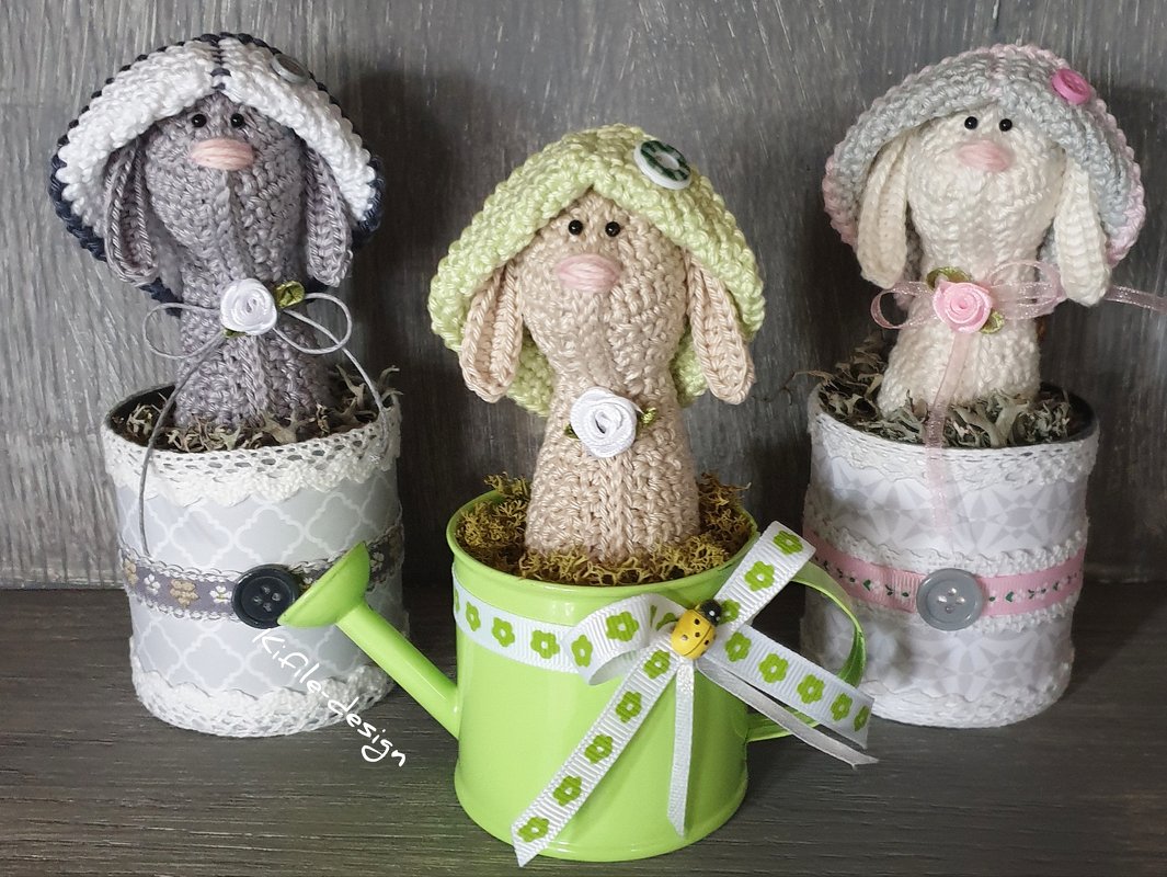 Drei gehäkelte Amigurumi-Hasen mit Hüten in dekorierten Töpfchen, mittlerer Hase in grünem Gießkännchen