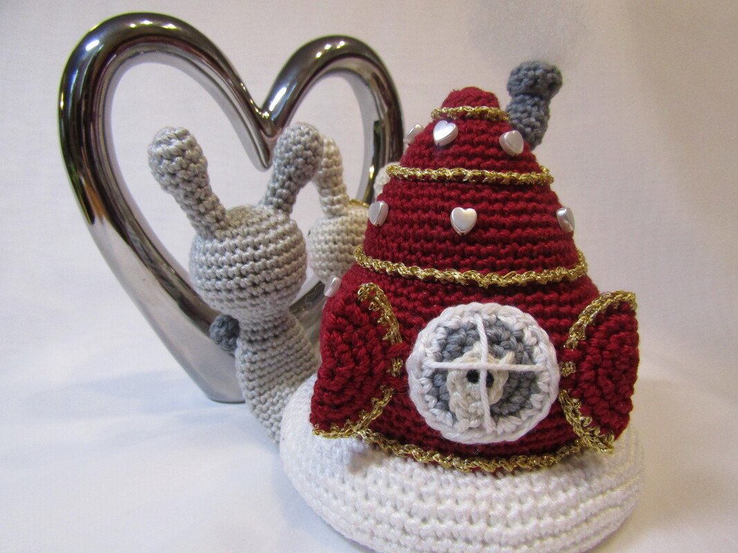 Nahaufnahme einer gehäkelten Amigurumi‑Schnecke mit roter Muschel, goldenen Borten und herzförmigen Perlen