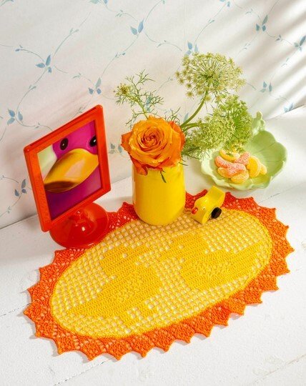 Ovales gehäkeltes Deckchen in Gelb und Orange mit gezacktem Rand, dekorativ arrangiert mit einer orangefarbenen Rose, Süßigkeiten und einem Bilderrahmen.